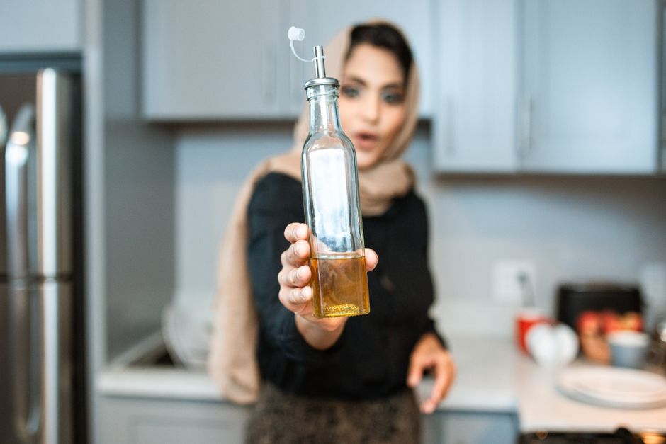 Positive ethnic woman wearing headscarf showing glass bottles of organic olive or sunflower oil while cooking in modern kitchen